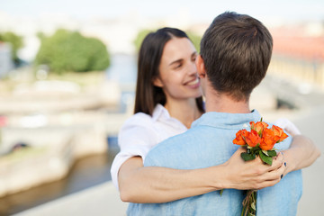 Naklejka premium Focus on small bouquet of orange roses in hands of girlfriend, happy affectionate woman falling in love with boyfriend cuddling him on roof