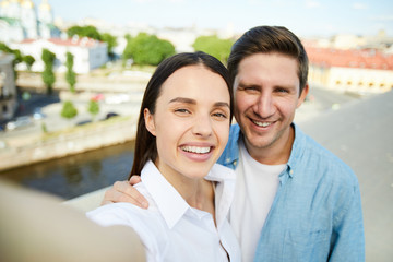Happy jolly young couple in casual shirts embracing each other and looking at camera while taking selfie on roof against cityscape