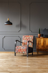 Colorful armchair with pattern standing in dark living room interior in real photo with lamp, molding on the wall and wooden cupboard with plant and decor