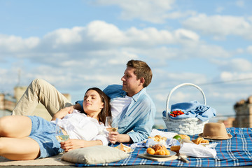 Carefree relaxed young couple in casual clothing lying on blanket while resting at roof picnic and drinking wine