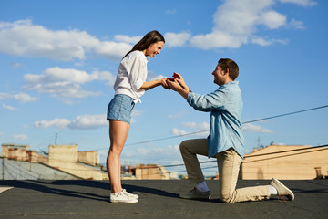 Happy loving man getting on one knee and opening jewelry box with engagement ring while making marriage proposal to beautiful woman on city roof