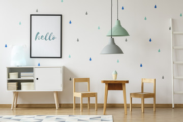 Poster above white cabinet next to wooden chairs and table in pastel kid's room interior. Real photo