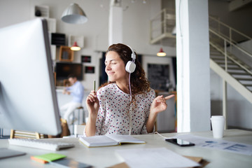 Joyous young businesswoman with headphones listening to dynamic music while working by desk in front of computer monitor - Powered by Adobe