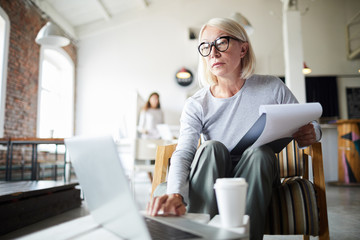 Mature businesswoman with document and laptop sitting on chair in office and browsing in the net