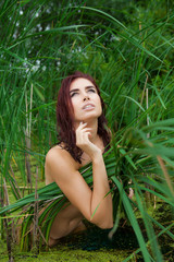 Fototapeta premium a naked young woman is standing on a swamp in a thicket of creepers with wet hair. Closed with grass