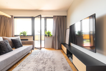 Television above countertop in spacious living room interior with grey sofa and balcony. Real photo