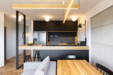 Lights above countertop of black kitchen in open space interior with sofa and table. Real photo