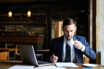 Serious handsome young bearded businessman drinking coffee and examining document while working in fashionable restaurant, he preparing report
