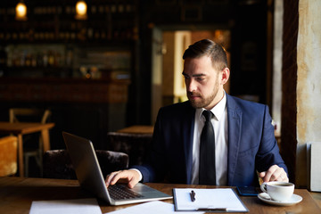 Fototapeta na wymiar Busy concentrated man in formal suit using laptop while checking file in cafe, he sitting at table and working in restaurant