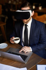 Serious young businessman in VR headset using laptop and interactive simulator for work while drinking coffee in restaurant