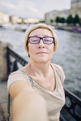 Senior female in glasses looking at camera while taking selfie on embankment of amazing city