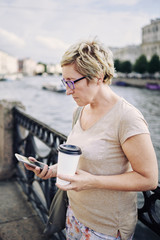 Senior lady in glasses standing on city street near river and using modern smartphone