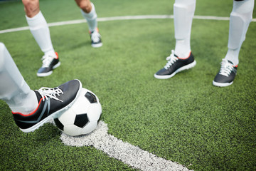 Foot of soccer player on ball lying on white line of football field with two men near by