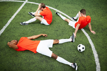 Tired or restful men in football uniform relaxing on green field during break between games