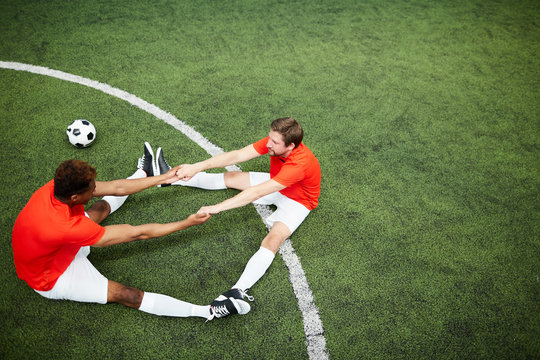 Two young footballers in uniform sitting on green field and holding by hands with their feet put against each other