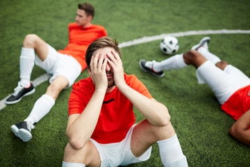Stressed or tired footballer sitting on green field and covering his face with hands on background of two mates