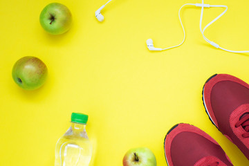Fitness flat lay. Healthy lifestyle and sport concept. Red sneakers, white earphones, apples and bottle of water on a yellow background.