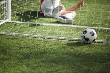 Fallen goalkeeper lying on green football field and soccer ball in gate