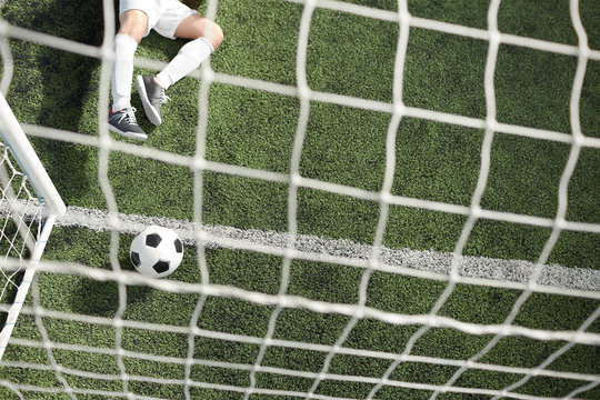 Overview of soccer ball in gates by net and legs of goalkeeper lying on green field