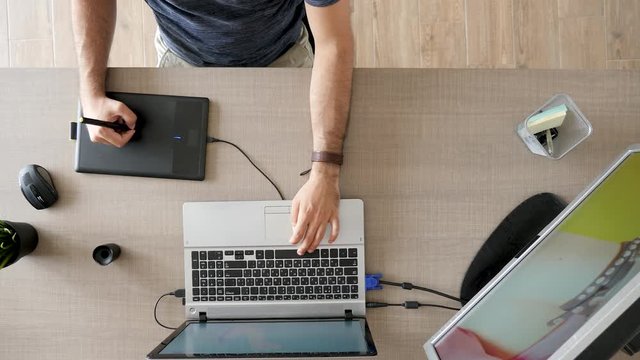 Graphic designer at his work desk drawing on digital tablet and his laptop is connected to an external monitor. Top view footage