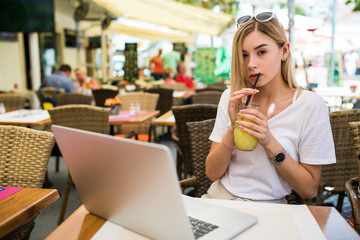 Young woman with glasses on her head smiling joyfully, resting at cafe and browsing internet using laptop computer, sitting at table with fruit shake people having lunch on background