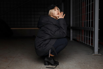 Young beautiful woman in fashion winter jacket and black stylish cap with fashionable pants with...