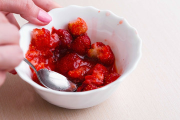 Woman kneads strawberries with a spoon and mixes it with sugar, making jam. Close-up hand.