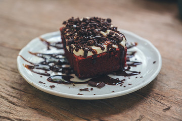 A piece of red velvet cake with cream and chocolate in white ceramic plate on wooden table