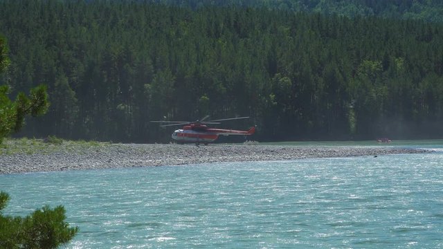 The Mil Mi-8 helicopter, which protects the forest from fires, standing on the bank of a mountain river, is preparing for take-off. A catamaran swims past with a group of people.