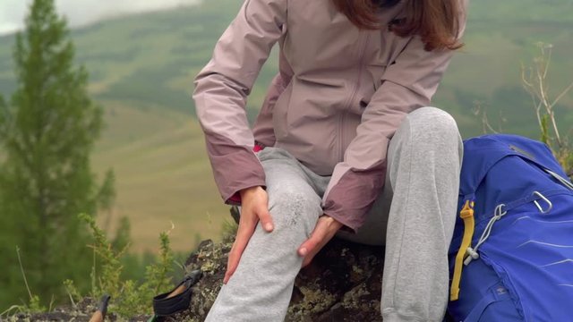 The girl was injured in the mountains. Tourists are provided with medical assistance. A woman in a hat holds on to a damaged leg sitting on a rock.