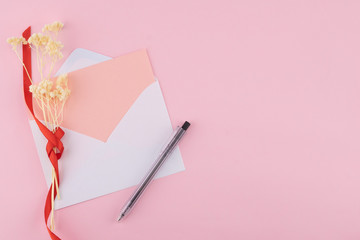 Blank pink card in white envelop and black pen with white dried flowers on pastel pink background with copy space