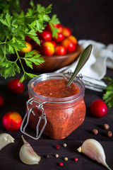 Tkemali sauce from red plums and ingredients for cooking garlic, parsley, dill, pepper with copy space on a dark wooden background