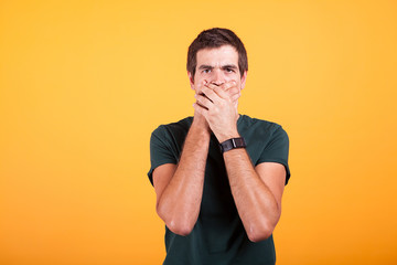 Attractive man in casual t-shirt covering his mouth in no talk sign on yellow background in studio...