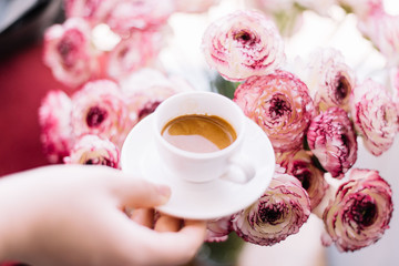 Hand holding a little cup of freshly brewed espresso coffee with a nice crema on the pink blossoming cappuccino Ranunculus flowers background, close up soft focus view
