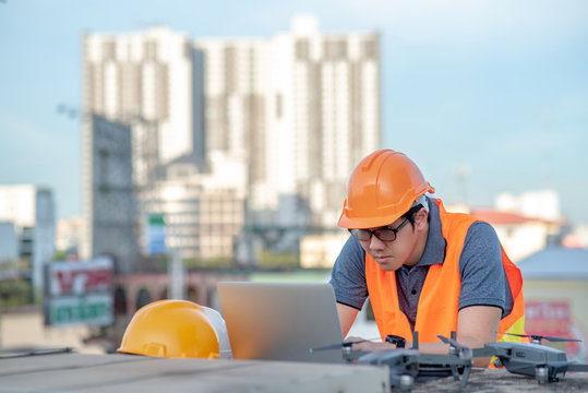 Young Asian man working with drone laptop and smartphone at construction site. Using unmanned aerial vehicle (UAV) for land and building site survey in civil engineering project.