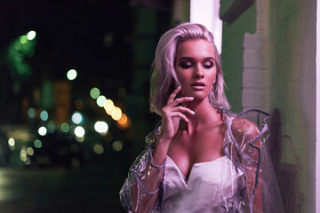 sensual young woman in transparent raincoat standing on street at night under pink light