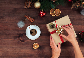 Christmas workplace top view. Girl’s hands on the wooden table holding Christmas present. Christmas wreath, decorations, gifts and ornaments.