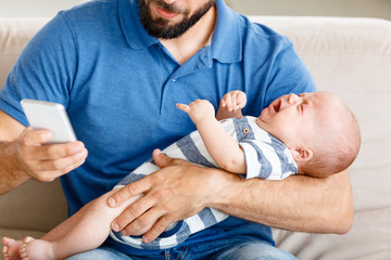Father holding crying baby boy and using smart phone