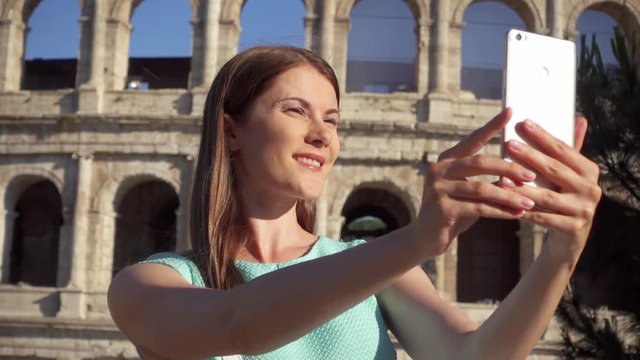 Woman do selfie on mobile phone near famous attraction Colosseum in Rome, Italy. Teenage girl smiling in slow motion. Happy female tourist enjoying her european vacation. Student travel through Europe