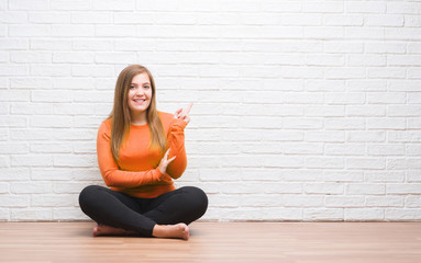 Young adult woman sitting on the floor in autumn over white brick wall very happy pointing with...