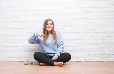 Young adult woman over white brick wall listening music wearing headphones with surprise face...