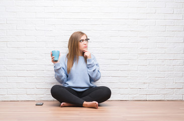 Young adult woman sitting on the floor over white brick wall driking cup of coffee serious face...