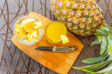 Top view of whole pineapple kept on a wooden table besides a glass filled with pineapple juice and a glass bowl with pineapple slices and a knife kept on the chopping board