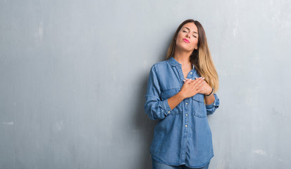 Young adult woman over grunge grey wall wearing denim outfit smiling with hands on chest with closed eyes and grateful gesture on face. Health concept.