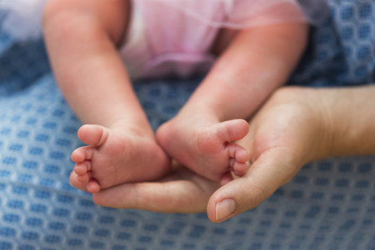 Ножки младенца, крохотной девочки в розовом боди, в руках мамы в голубом платье  