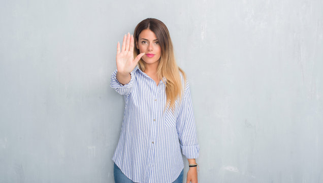 Young adult woman over grey grunge wall wearing fashion business outfit doing stop sing with palm of the hand. Warning expression with negative and serious gesture on the face.