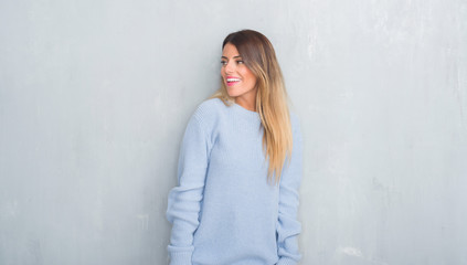 Fototapeta premium Young adult woman over grey grunge wall wearing winter outfit looking away to side with smile on face, natural expression. Laughing confident.