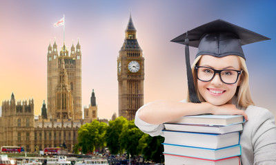 Obraz premium education, school, knowledge and graduation concept - happy graduate student girl in mortarboard with stack of books over houses of parliament in london background