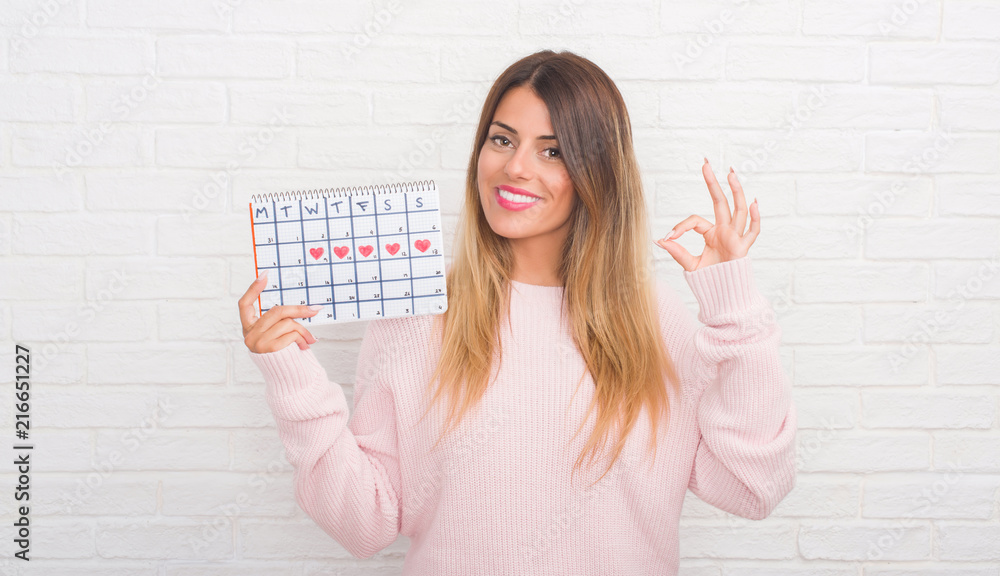 Sticker young adult woman over white brick wall holding period calendar doing ok sign with fingers, excellen