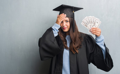 Young brunette woman over grunge grey wall wearing graduate uniform holding dollars stressed with...